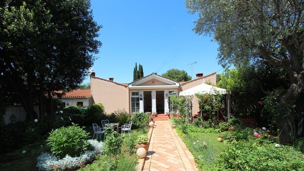 Un sentier de jardin mène à un bâtiment doté d’une entrée centrale, qui est entouré d’une végétation luxuriante et de plantes en fleurs.