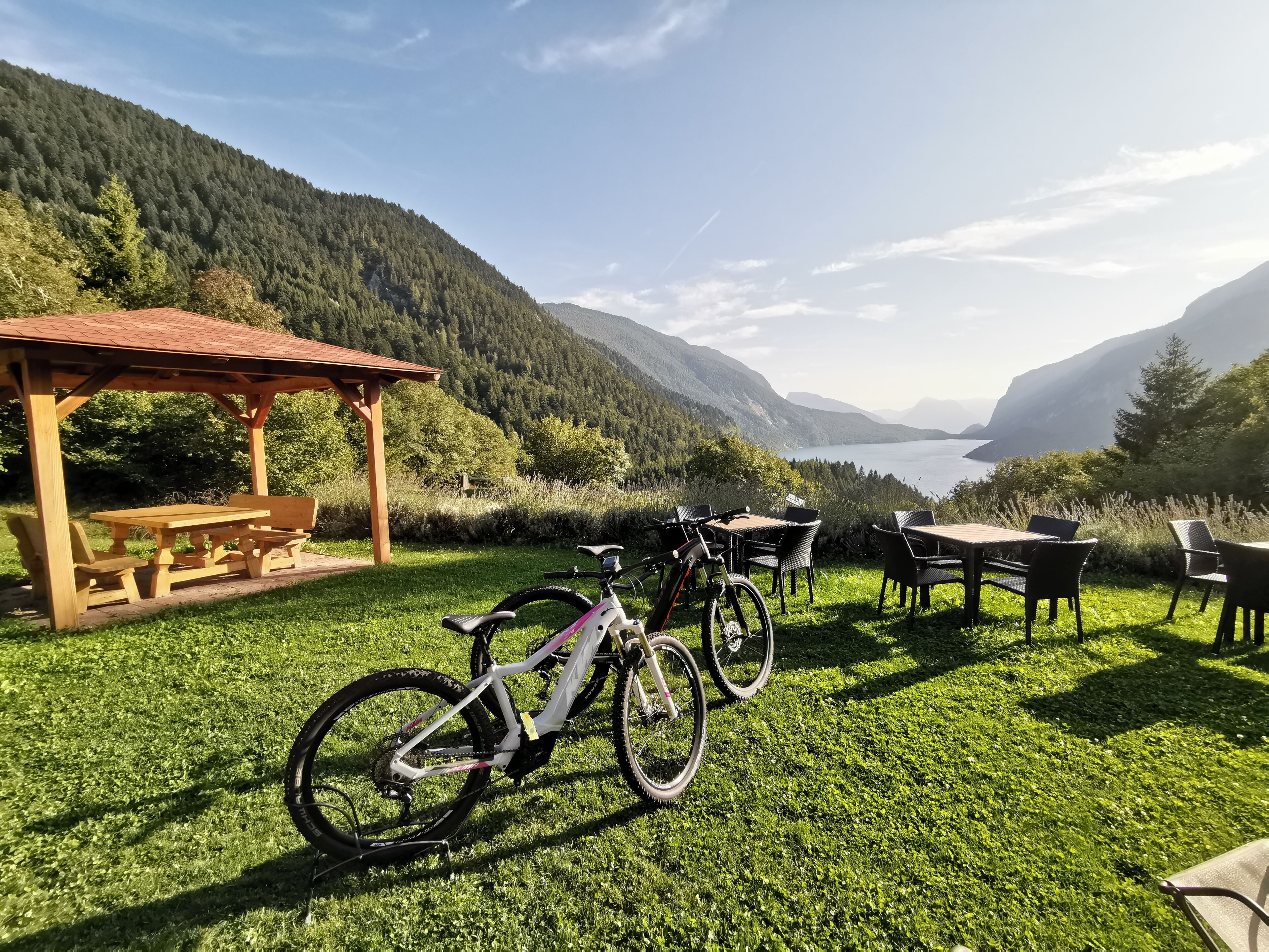 Zona de picnic amb una pèrgola de fusta i taules, una bicicleta, i vista d'un llac i muntanyes.