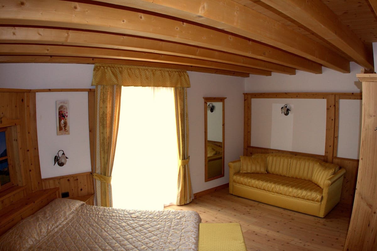 Een kamer met een bed, een gele bank, houten balken in het plafond en een raam met gordijnen.