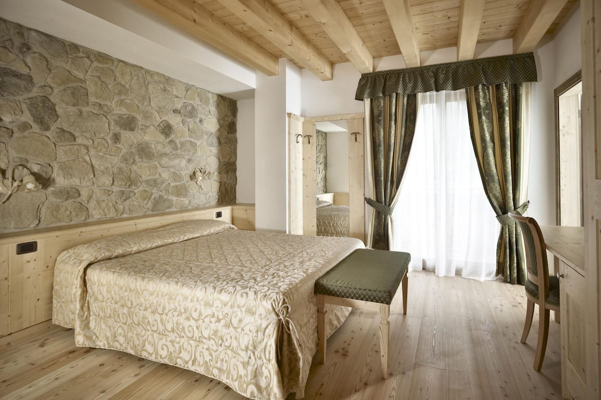 Een slaapkamer met een stenen muur, houten balken in het plafond, een bed met een geborduurde spreideken, een bankje en gordijnen.