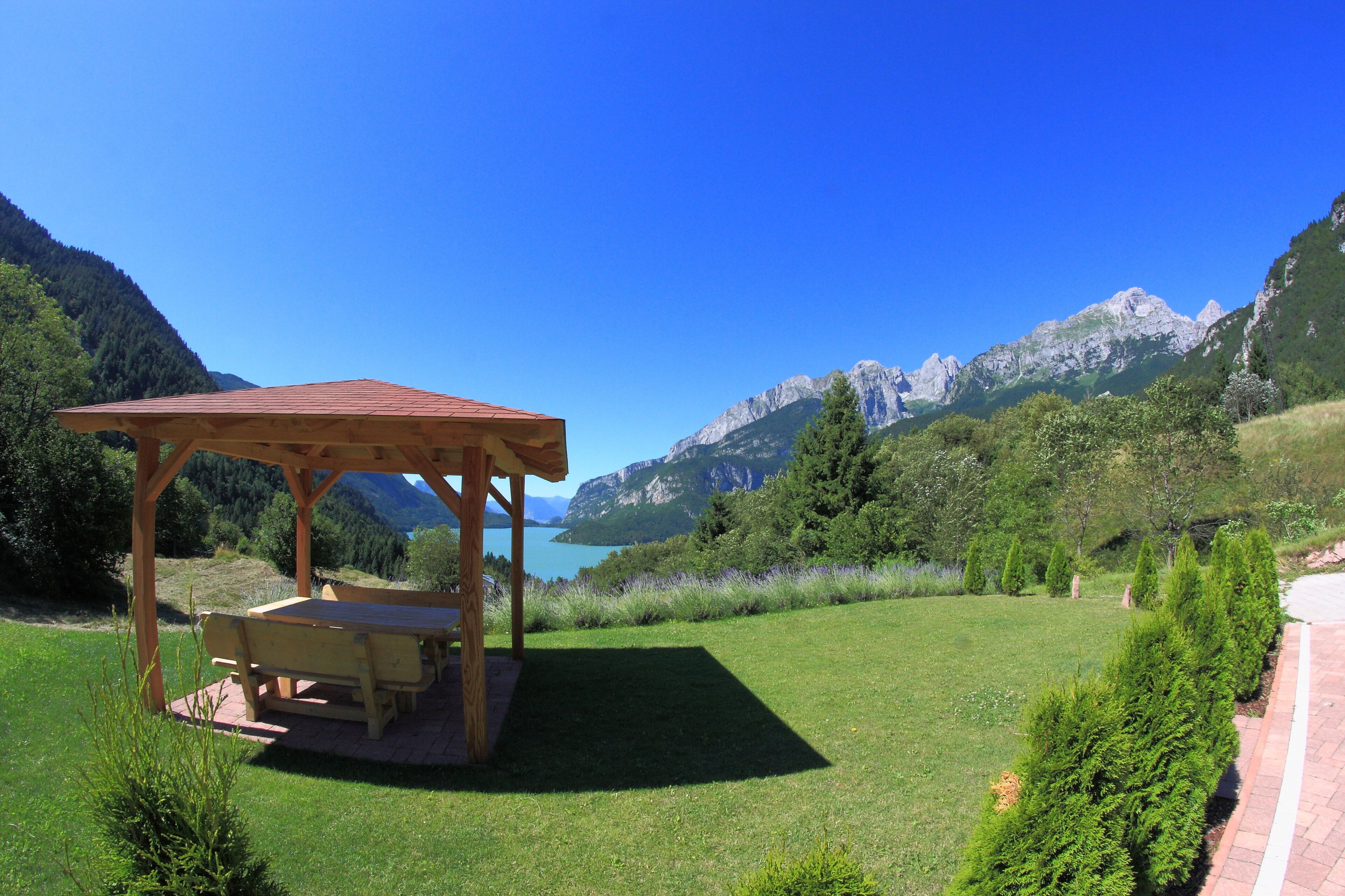 A wooden gazebo with a bench, surrounded by a well-maintained lawn and a view of mountains and a lake.