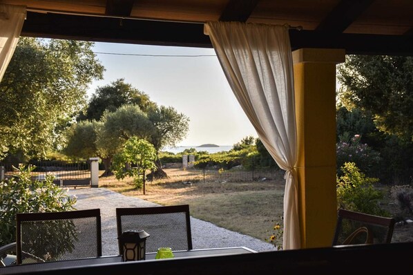 An outdoor patio with a table and chairs, overlooking a garden with trees and a view of the sea.