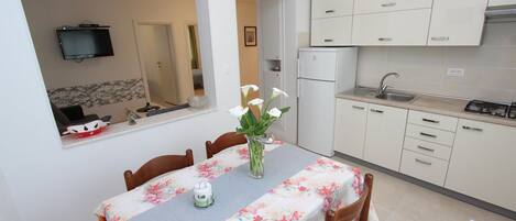 Comedor con una mesa preparada para cuatro personas, un jarrón con flores y una cocina con armarios blancos y un refrigerador.