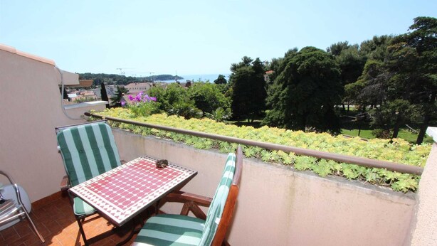 A balcony with a table and two striped chairs, overlooking a garden and distant buildings.