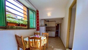Un comedor con una mesa redonda y sillas de madera, una cocina al fondo y una ventana con persianas.