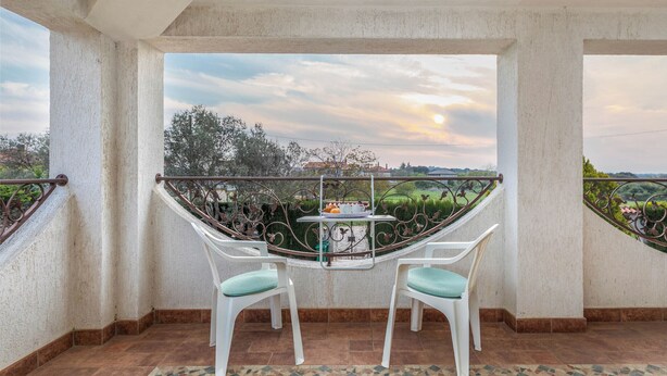 A balcony with two white chairs, a small table with a tray of items, and a view of a landscape with trees and a sunset. - VILLA NERA Three-Bedroom Apartment (Pula)