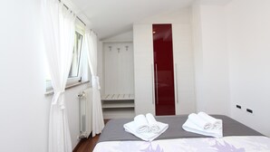 Une chambre d’hôtel avec un lit, une fenêtre avec des rideaux blancs, un radiateur, une porte rouge et une armoire blanche.