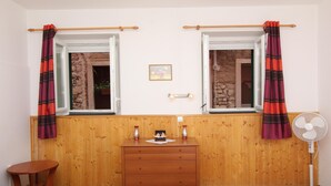 A room with wooden paneling, a wooden dresser, two windows with curtains, a small table, and a standing fan. - PUPA Studio Apartment (Rovinj)