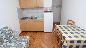 A compact kitchen with wooden cabinets, a sink, and a white refrigerator. There's a dining table with a checkered tablecloth and a wooden chair. - LORA One-Bedroom Apartment 3 (Rovinj)