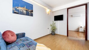 A living room with a sofa, a potted plant, a flat-screen TV, and a painting of a coastal town. - SILVANA One-Bedroom Apartment with Terrace 2 (donji) (Rovinj)