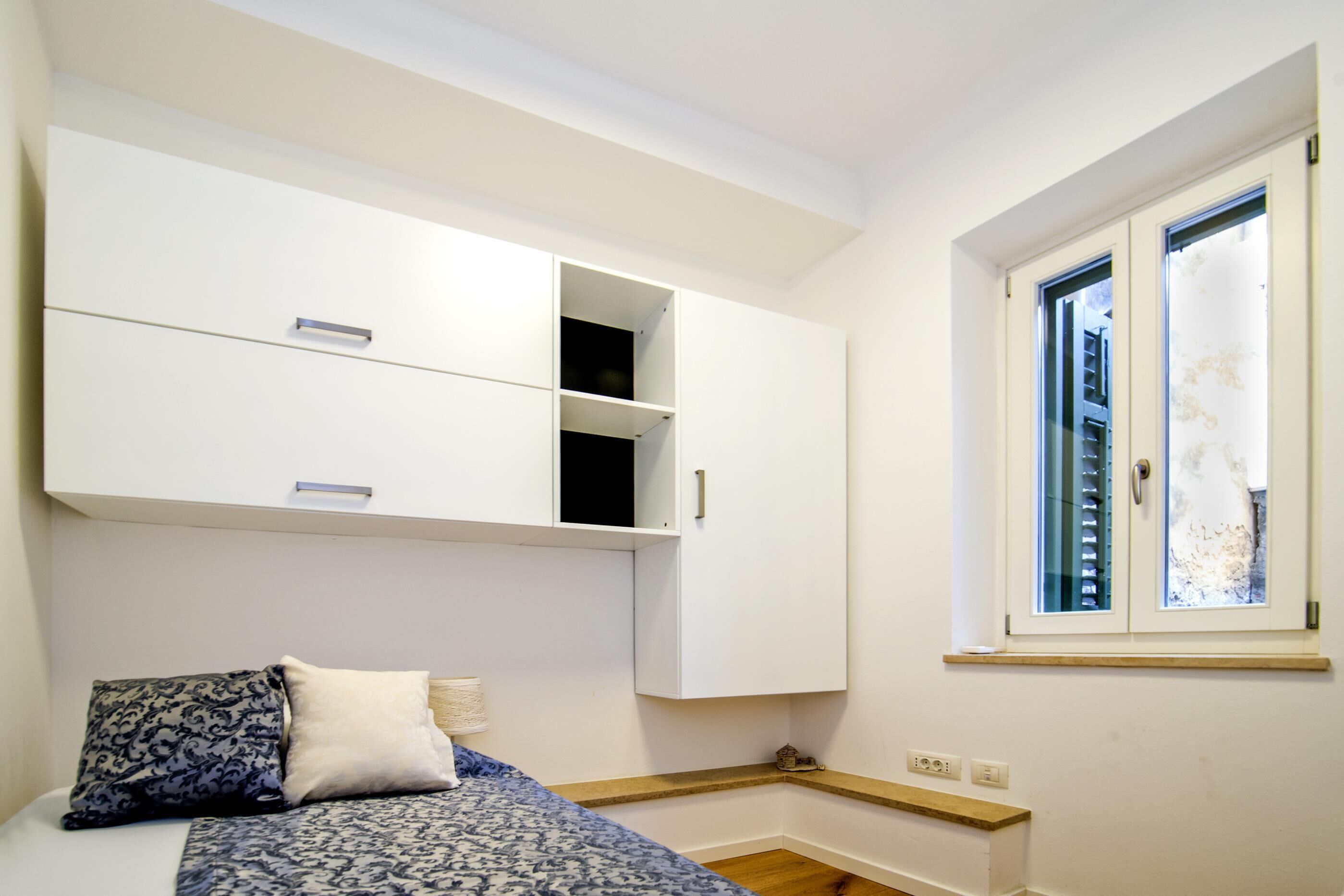 A bedroom with a bed, a wardrobe, a window, and a small shelf.