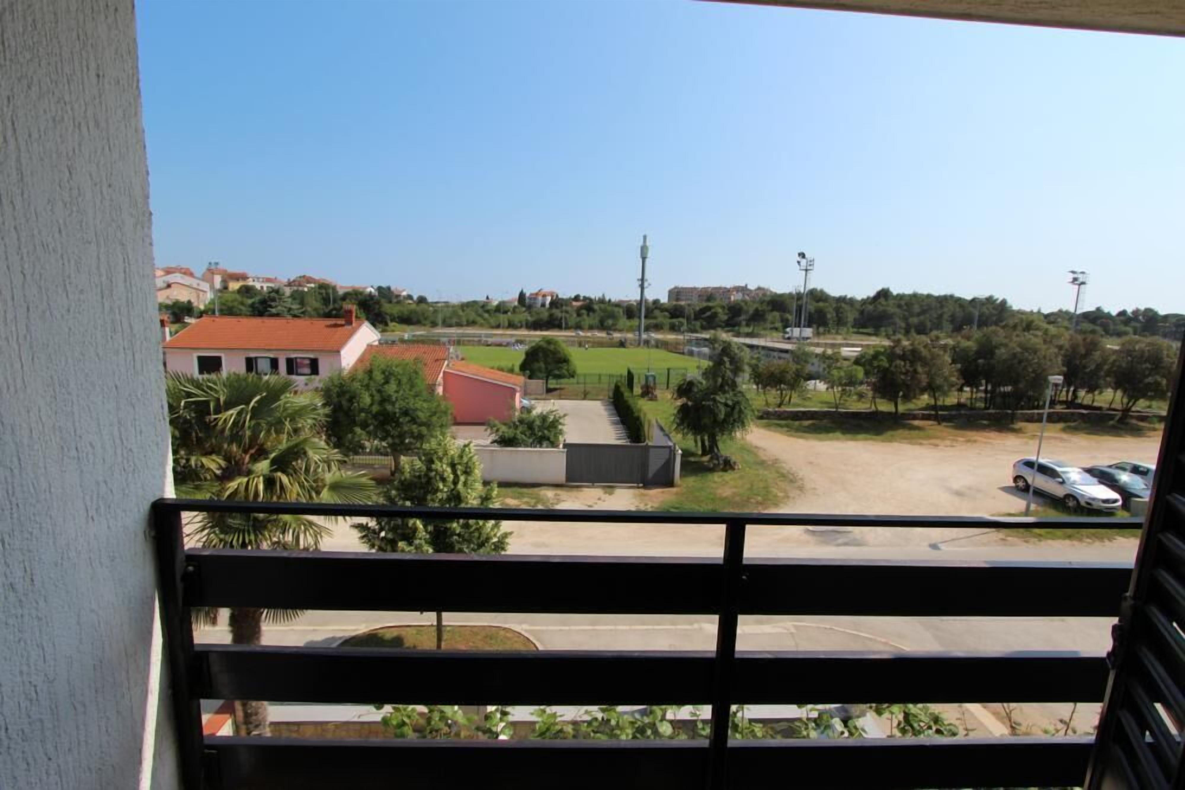 A balcony with a view of a parking lot, a sports field, and surrounding buildings.