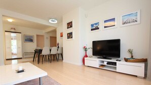 A modern living room with a flat-screen TV on a white entertainment unit, a dining area with a table and chairs, and a hallway leading to another room.