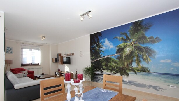 A living room with a large beach mural, a dining table with candles, and a sofa with red cushions. - DADO One-Bedroom Apartment (Putini)