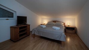 Ein Schlafzimmer mit einem Bett, zwei Nachttischen mit Lampen, einem Fernseher auf einem Holzständer und einem Fenster mit Blick zum Himmel.