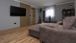 A living room with a large sectional sofa, a flat-screen TV mounted on the wall, a wooden floor, and a guitar leaning against the wall. - ENI One-Bedroom Apartment 1 (mali) (Rovinj)