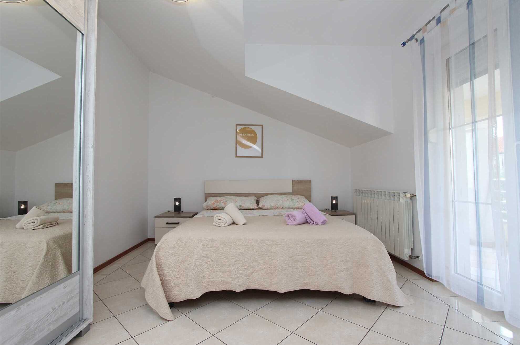 Un dormitorio con una cama grande, una mesita de noche, una lámpara de noche, un cuadro en la pared y una ventana con cortinas.