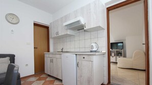 Eine kompakte Küche mit integriertem Geschirrspüler, Mikrowelle und Herd. Es gibt eine kleine Sitzecke mit Sofa und Sessel. Im angrenzenden Wohnzimmer ist ein Fernseher an der Wand montiert.