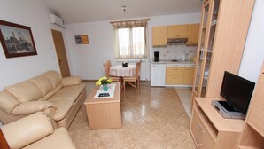 A living room with a beige sofa, a coffee table with a vase, a dining table with chairs, a kitchen area with cabinets, and a television on a wooden stand. - LILIANA One-Bedroom Apartment (Rovinjsko Selo)