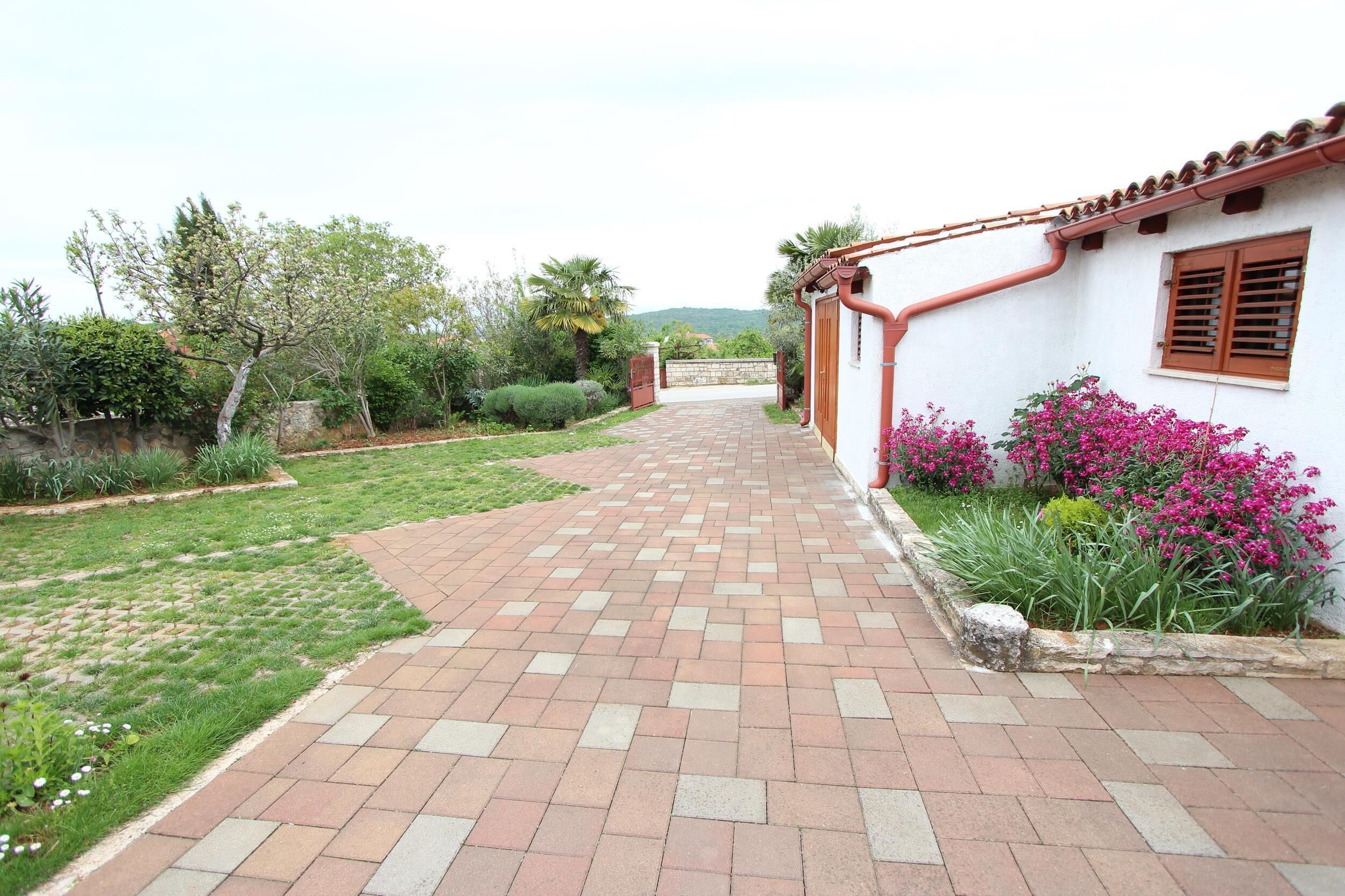 Un camino pavimentado que conduce a un edificio blanco con techo rojo, rodeado de vegetación y plantas en flor.