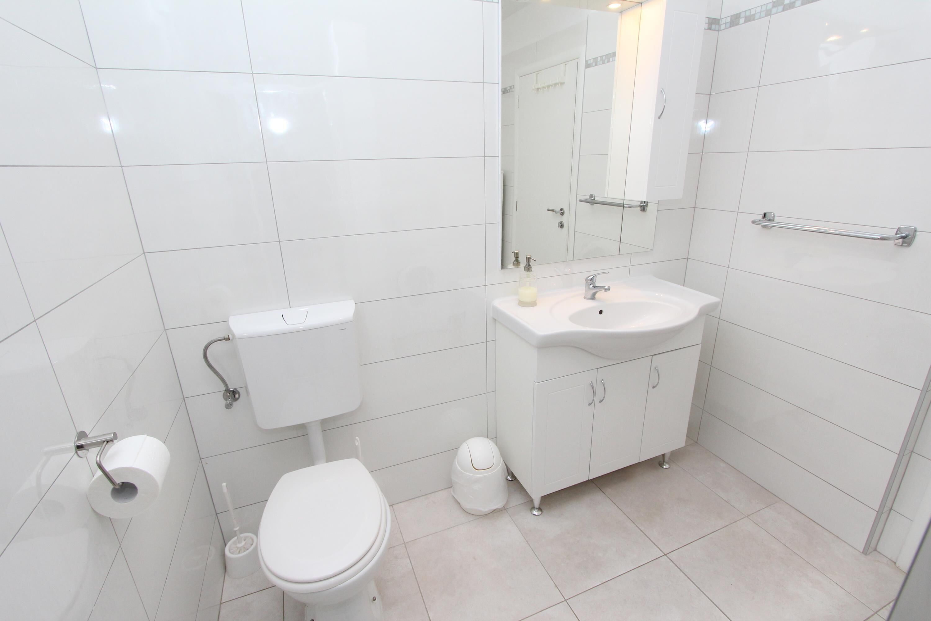 A modern bathroom with white tiles, a toilet, a sink with a mirror, and a handrail on the wall.