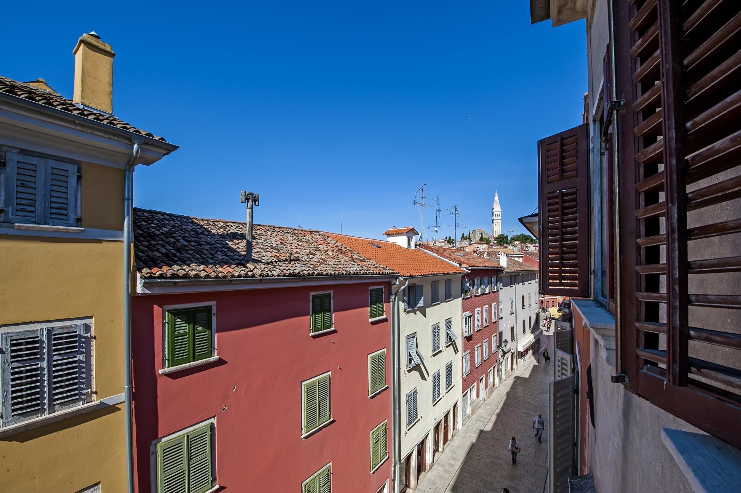 A narrow street with colorful buildings, a prominent tower in the background, and a clear blue sky.