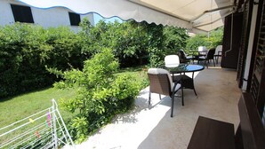 An outdoor patio area with a covered seating area, surrounded by greenery and a white building with a blue trim. - APOLLONIO Studio Apartment with Terrace (Rovinj)