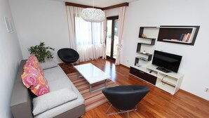 A modern living room with a grey sofa, black armchair, and a white coffee table. There is a TV on a white stand, and a bookshelf with books and decorative items. - MITTON 2 Two-Bedroom Apartment (Motovunska) (Rovinj)
