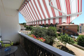 Un balcón con toldo a rayas, una mesa con superficie de vidrio y vistas a edificios y zonas verdes.