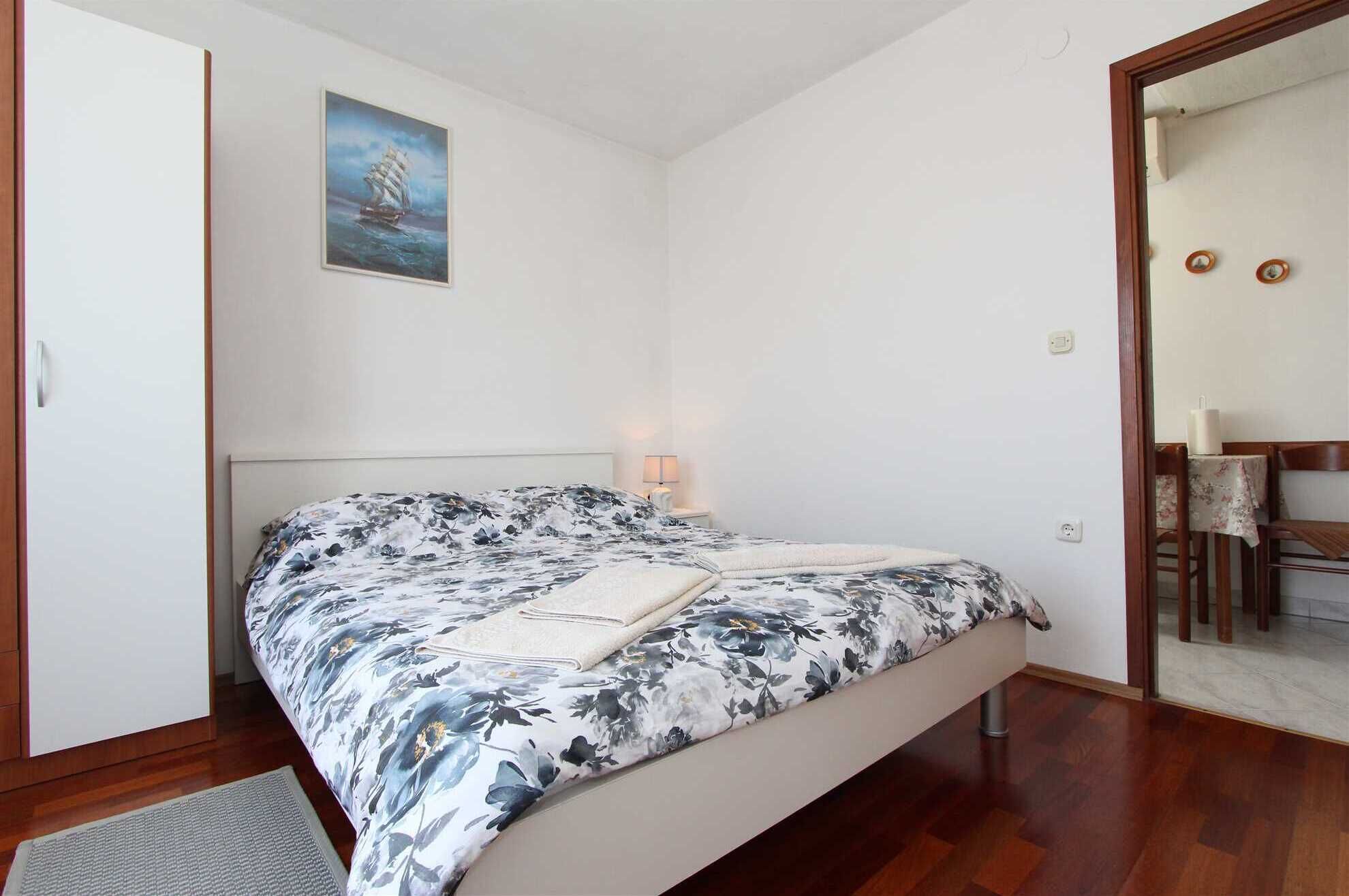 Ein Schlafzimmer mit Bett, Nachttisch, Lampe, einem Bild an der Wand und einer Tür zu einem anderen Raum.