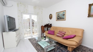 Un salón con sofá beige, una mesa de centro de vidrio, un mueble blanco y un televisor sobre un pedestal.
