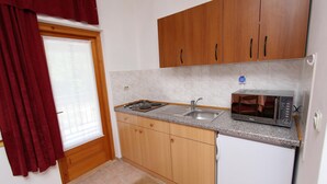 A compact kitchen with wooden cabinets, a microwave, a sink, and a window with red curtains. - MARIZA Studio with Balcony (Rovinj)