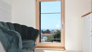 Una habitación con un sillón azul turquesa, un armario de madera y un ventanal corredera con vistas a casas y al mar.