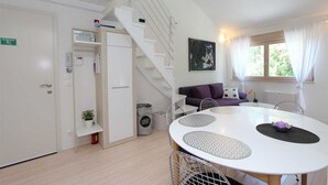 Una habitación moderna con una escalera blanca, un sofá morado, una mesa redonda con superficie blanca y una ventana con cortinas blancas.