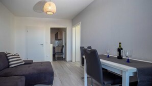 Een moderne woonkamer met een grijze bank, een afstandsbediening op een bijzettafeltje, een eethoek met een witte tafel, twee stoelen en een fles wijn.