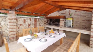 Een terras met eethoek, een stenen muur, een open haard van baksteen, houten balken en een tafel gedekt voor vier personen.