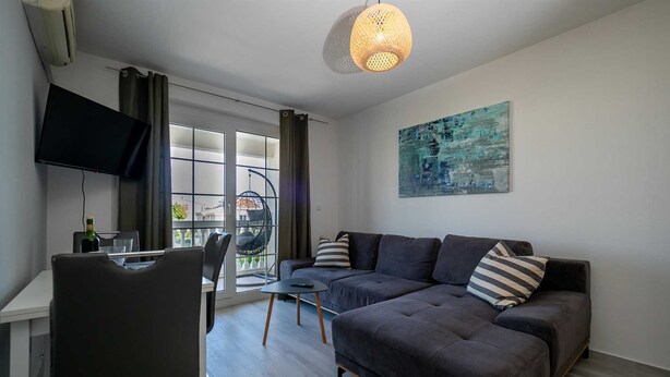 Een moderne woonkamer met een donkergrijze bank, een hangstoel, een eettafel met stoelen, een flatscreen-televisie en een groot raam met gordijnen.
