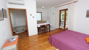 Un dormitorio con cama, escritorio y silla, una pequeña kitchenette y un balcón con cortinas.