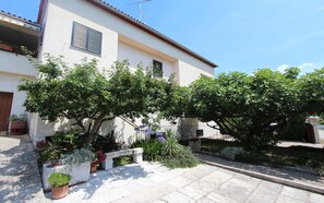 A building with a white facade, a balcony with a striped awning, and a garden area with various potted plants and a bench. - MITTON One-Bedroom Apartment (Cademia) (Rovinj)