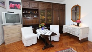 Ein Zimmer mit einem Holzschrank, einem Fernseher, einem Tisch mit weißer Spitzenleinentuch und zwei weißen Stühlen. An der Wand hängt ein Spiegel.