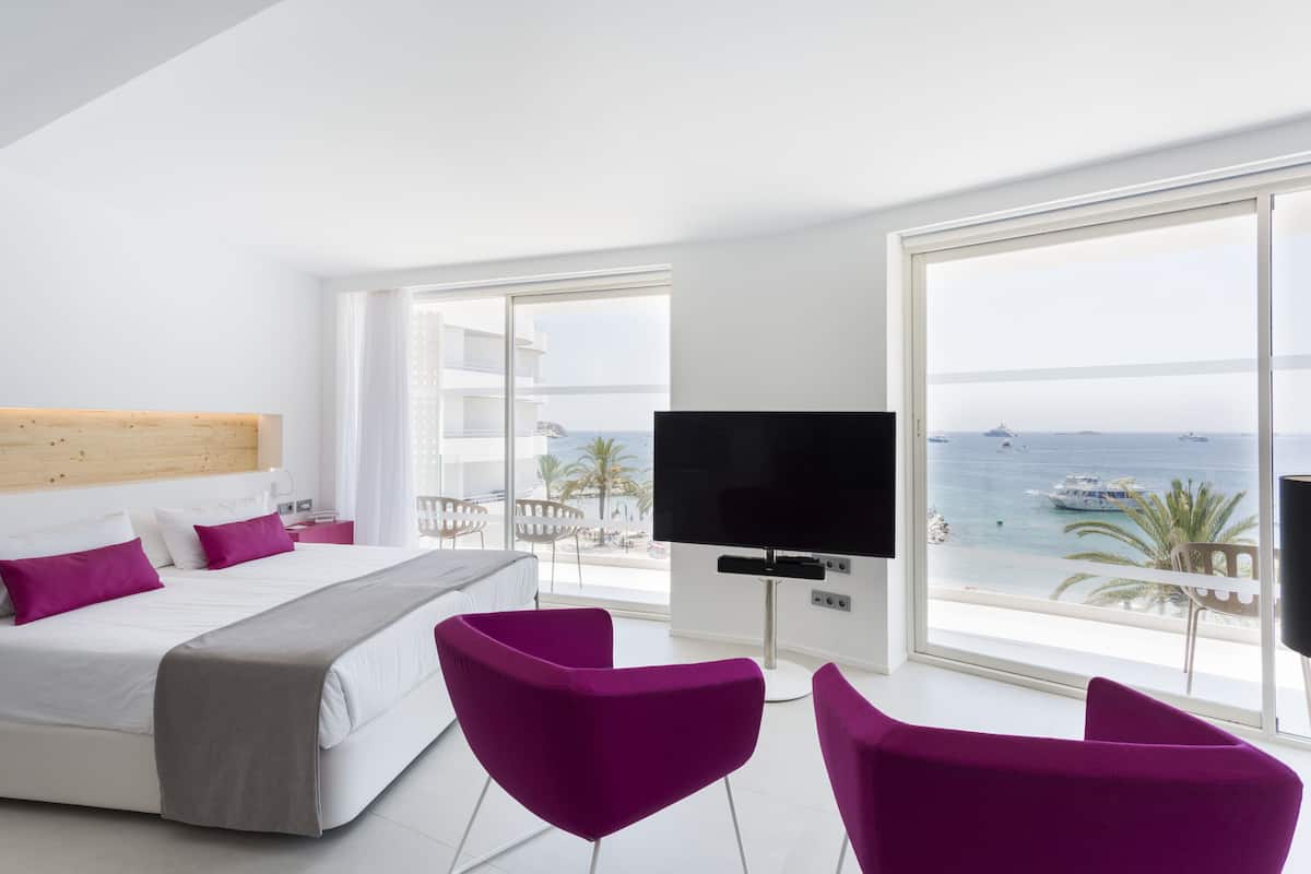 Een moderne hotelkamer met een groot bed, een flatscreen-tv en een balkon met uitzicht op zee en boten.