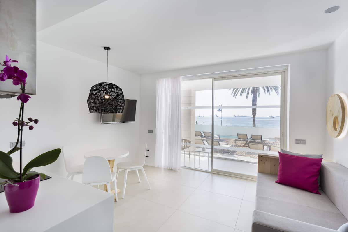 Een moderne woonkamer met een bank, een eettafel en stoelen. Er is een groot raam met uitzicht op het strand en palmbomen.
