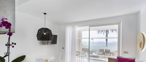 A modern living room with a sofa, a dining table, and chairs. There is a large window with a view of the beach and palm trees.