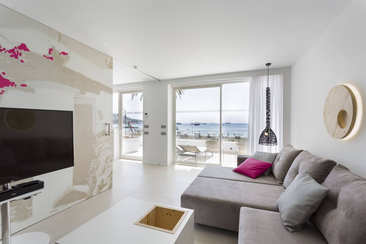 Een moderne woonkamer met een grote televisie, een bank met kussens, een salontafel en uitzicht op het strand door grote ramen.