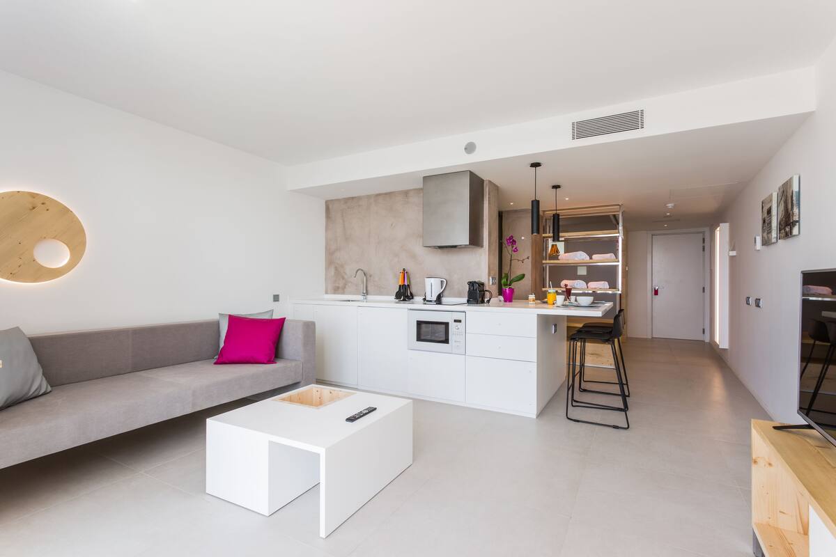 Een moderne keuken met witte kasten, een inbouwoven en een koffieapparaat. Er is een grijze bank met een roze kussen, een witte salontafel, en een houten bankje met krukken.