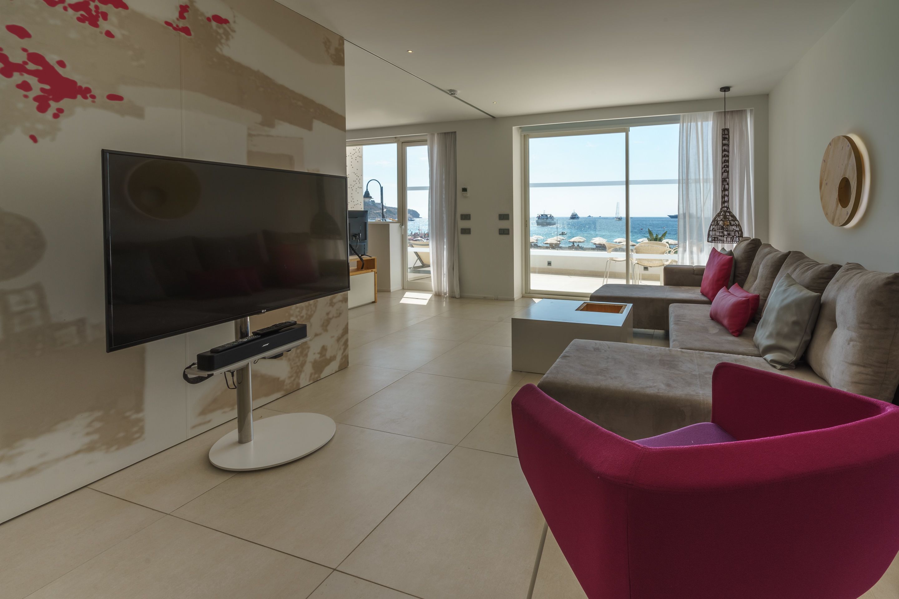 A modern living room with a flat-screen TV, a pink chair, a sofa with cushions, and a large window offering a view of the sea.