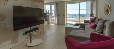 A modern living room with a flat-screen TV, a pink chair, a sofa with cushions, and a large window offering a view of the sea.