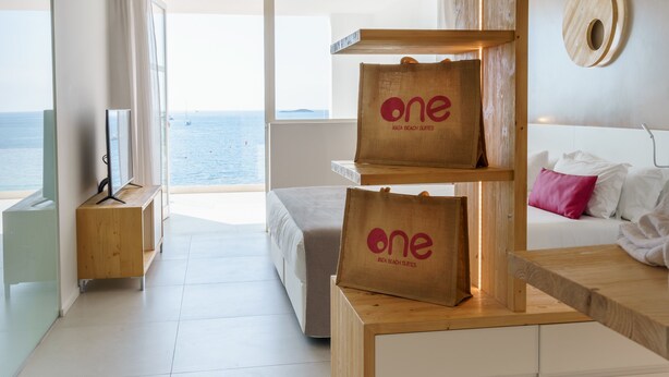 A modern hotel room with a large window offering a view of the sea, a wooden desk with a 'one' branded bag, a bed with white bedding, and a minimalist decor. - One Ibiza Suites (Ibiza Town)