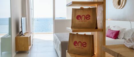 A modern hotel room with a large window offering a view of the sea, a wooden desk with a 'one' branded bag, a bed with white bedding, and a minimalist decor.