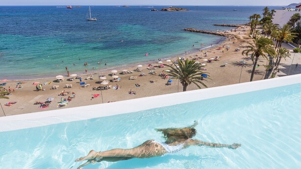 A pool with a person swimming, a beach with umbrellas and people, and a clear view of the sea. - One Ibiza Suites (Ibiza Town)